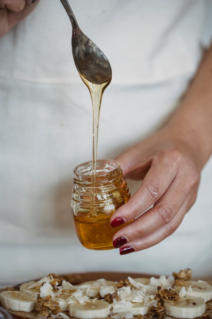 A hand drizzling honey from a jar over fresh banana slices and granola for a sweet treat.